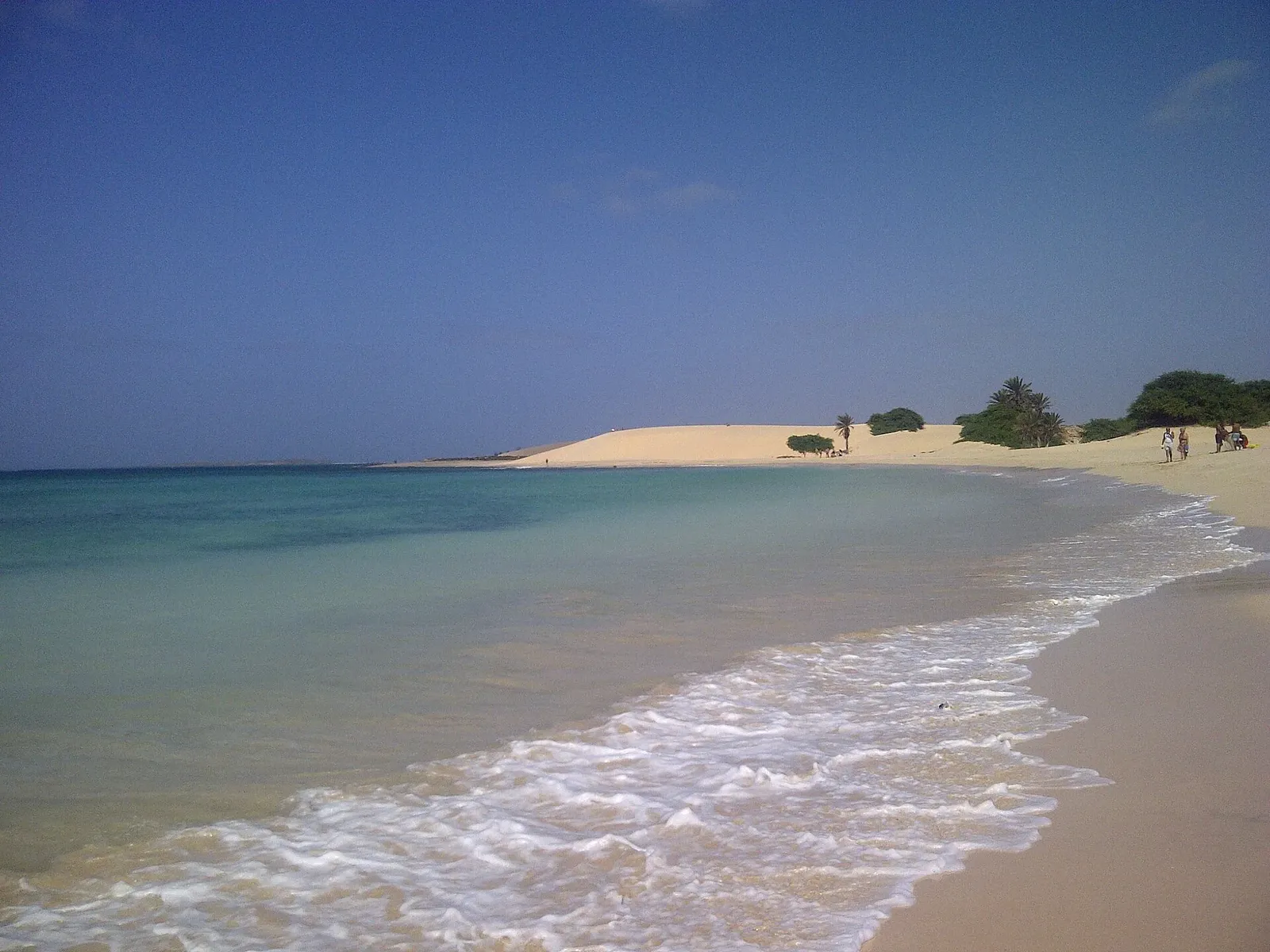 Plage de sable doré bordée d'une mer turquoise, avec des vagues et une grande dune au fond sous un ciel bleu