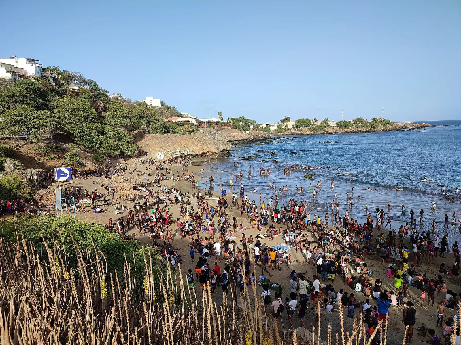 Plage de sable bondée de baigneurs dans une petite baie rocheuse, avec une végétation et des maisons blanches sur la colline