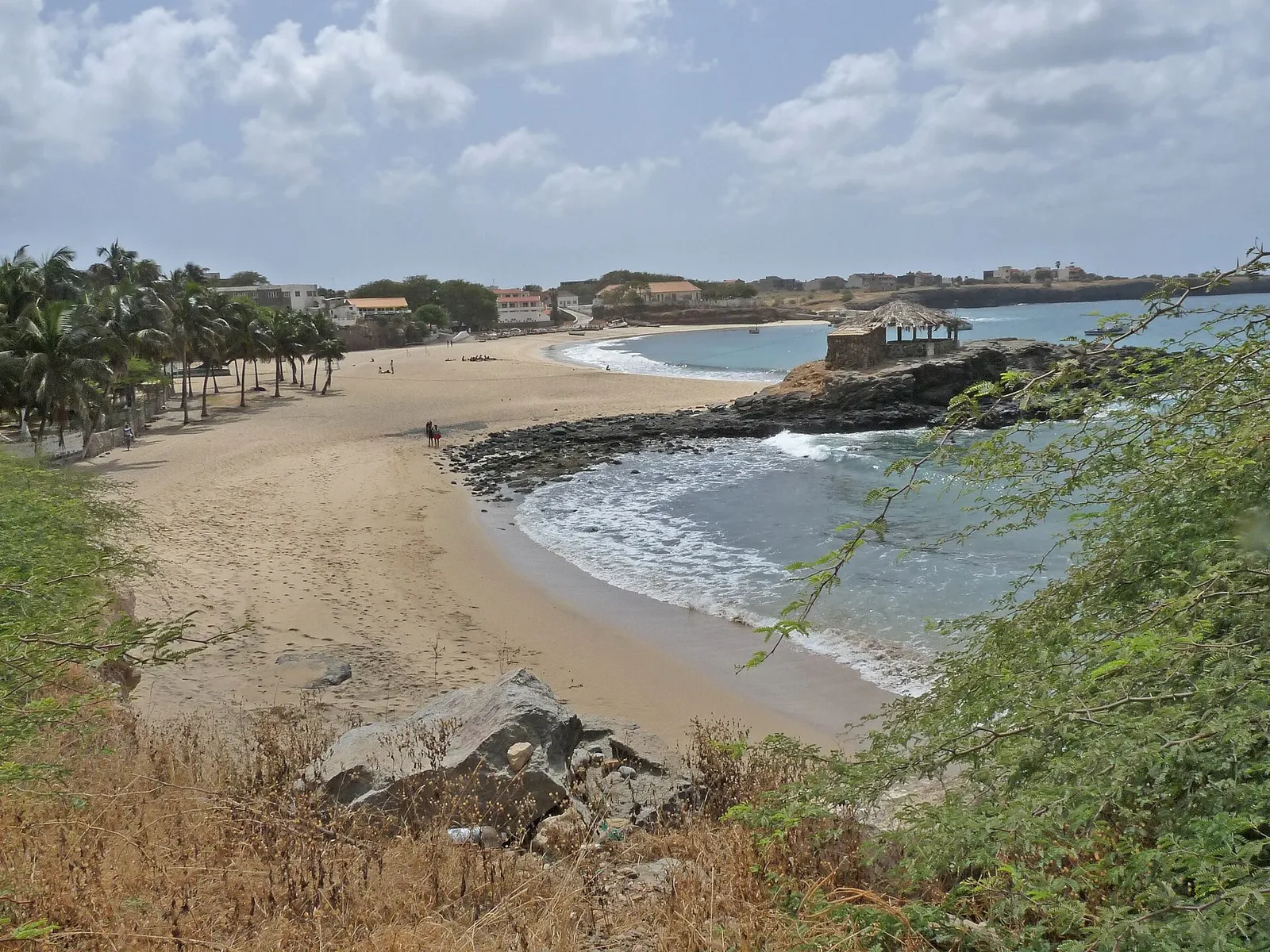Plage de sable bordée de palmiers, avec des rochers, une paillote sur un promontoire et des maisons au loin