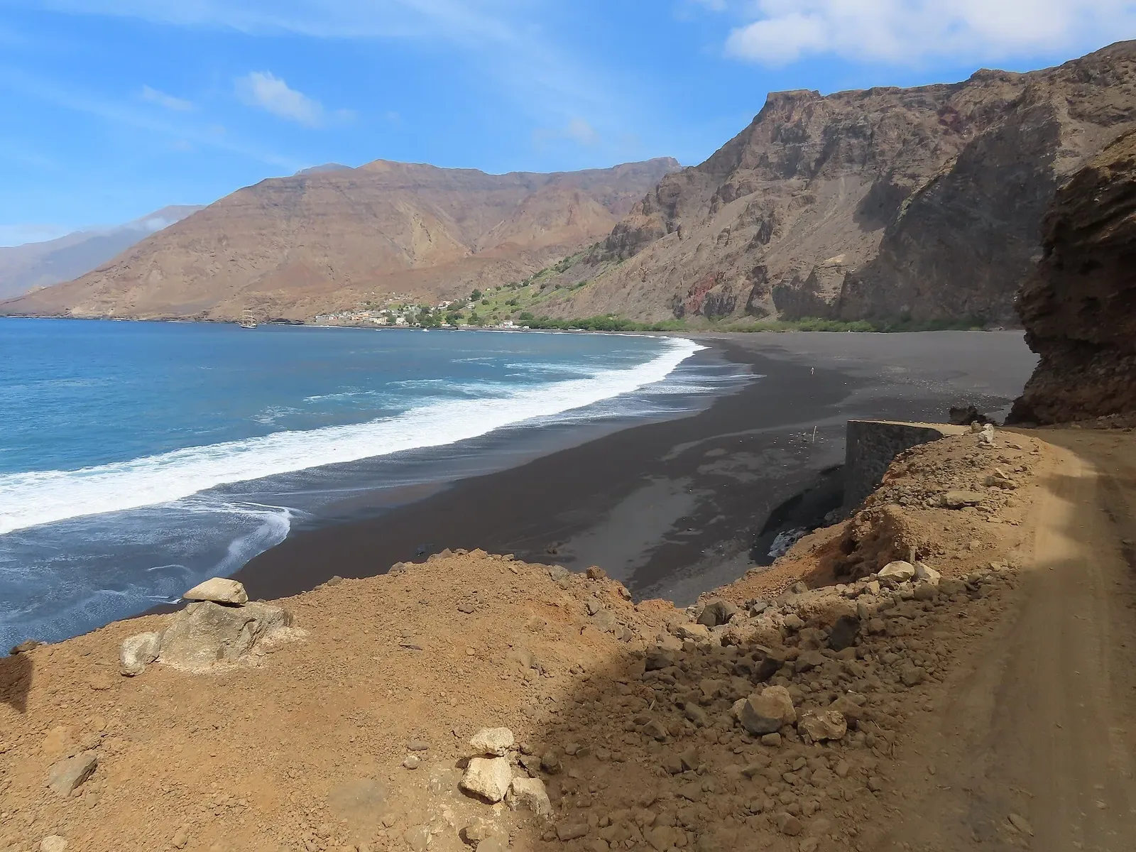 Plage de sable noir battue par les vagues, au pied de montagnes brunes, avec un village au loin et une piste de terre au premier plan