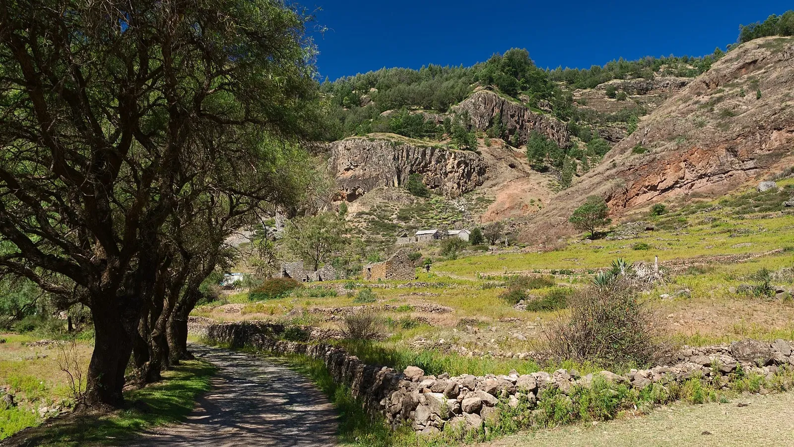 Sentier pavé longeant un muret de pierres sèches, dans une vallée verte dominée par des falaises rousses sous un ciel bleu