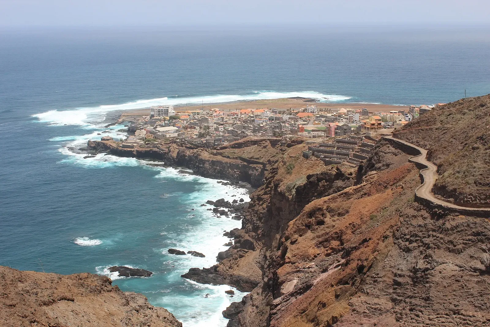 Village côtier vu depuis les falaises volcaniques, avec l'océan et ses vagues et une route en corniche à droite