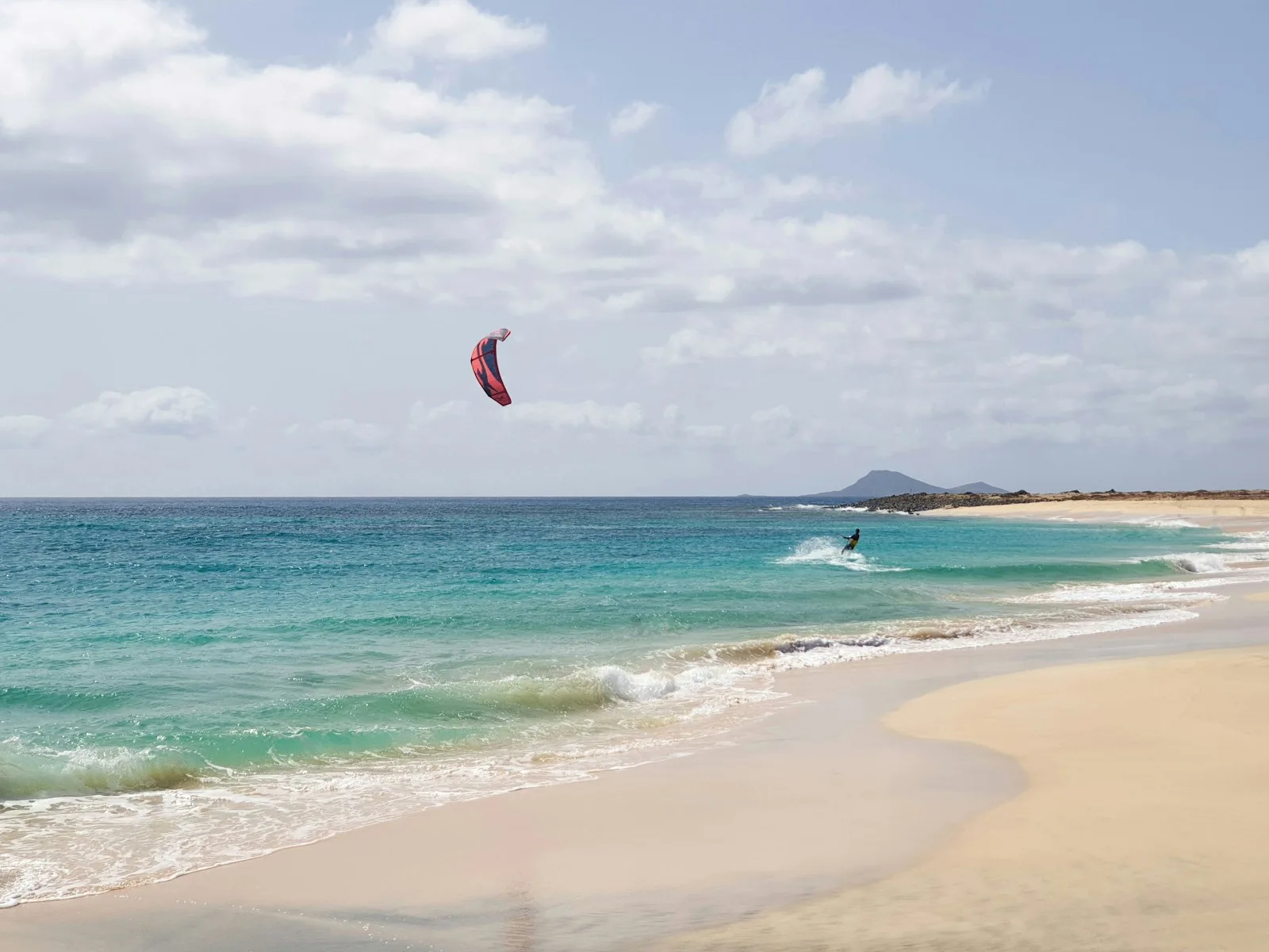 Kitesurfeur avec une aile rouge dans les vagues turquoise, devant une plage de sable doré et une colline au loin